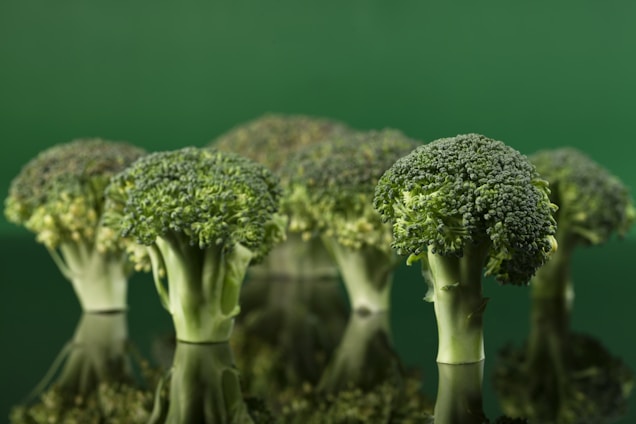 Several broccoli heads are standing upright on a reflective surface, creating a mirrored effect. The background is a solid, vibrant green, enhancing the fresh appearance of the broccoli.