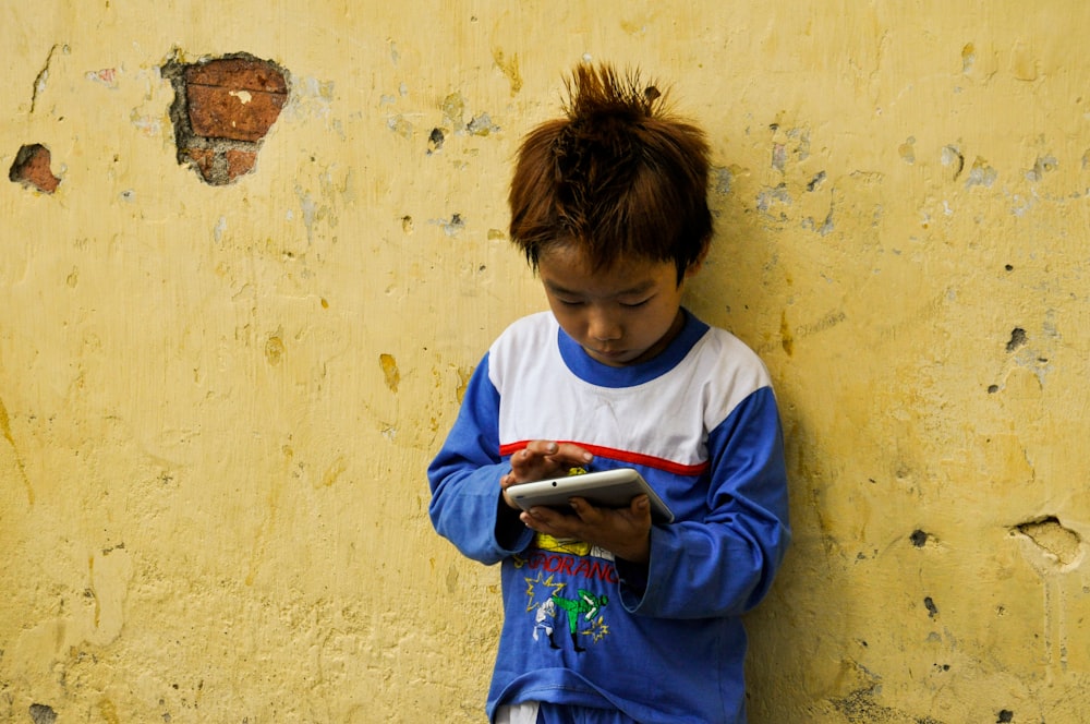 a young boy standing against a yellow wall looking at a tablet