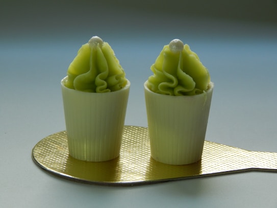 Two small, decorative dessert cups with ridged exteriors and light green frosting topped with a white pearl-like decoration. They are placed on a textured, gold-colored surface.