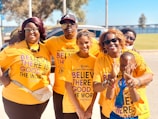 A group of six people stand close together, smiling at the camera. They are all wearing matching orange T-shirts that carry a positive message about believing in the good in the world. The setting appears to be outdoors, with trees and a clear blue sky in the background.