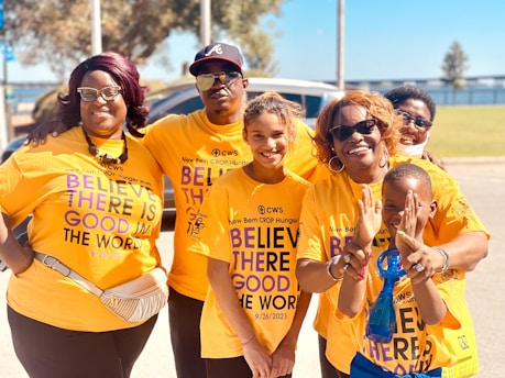 A group of six people stand close together, smiling at the camera. They are all wearing matching orange T-shirts that carry a positive message about believing in the good in the world. The setting appears to be outdoors, with trees and a clear blue sky in the background.