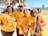 A group of six people stand close together, smiling at the camera. They are all wearing matching orange T-shirts that carry a positive message about believing in the good in the world. The setting appears to be outdoors, with trees and a clear blue sky in the background.