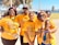 A group of six people stand close together, smiling at the camera. They are all wearing matching orange T-shirts that carry a positive message about believing in the good in the world. The setting appears to be outdoors, with trees and a clear blue sky in the background.