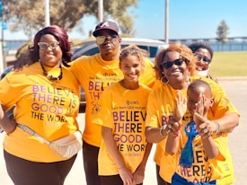 A group of six people stand close together, smiling at the camera. They are all wearing matching orange T-shirts that carry a positive message about believing in the good in the world. The setting appears to be outdoors, with trees and a clear blue sky in the background.