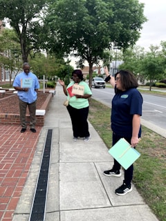 Three people are standing on a sidewalk next to a street, under a group of trees. Two of them are holding signs that read 'North Carolina' and 'New Bern,' while the third person is gesturing with their hand. The background includes a patch of grass, trees, and a road with parked cars.