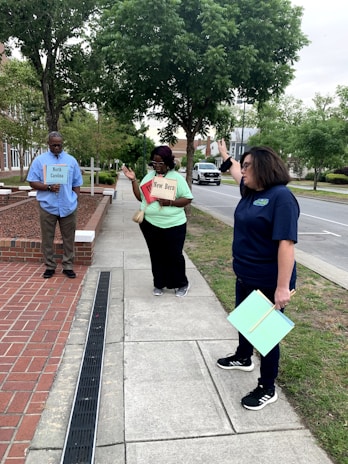 Three people are standing on a sidewalk next to a street, under a group of trees. Two of them are holding signs that read 'North Carolina' and 'New Bern,' while the third person is gesturing with their hand. The background includes a patch of grass, trees, and a road with parked cars.