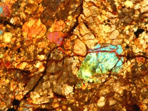 A detailed close-up of a rock or mineral surface, showcasing a vibrant mix of colors and intricate crystalline patterns. The surface features a rich array of gold, orange, and copper hues interspersed with bright patches of blue and green, giving it an iridescent quality.