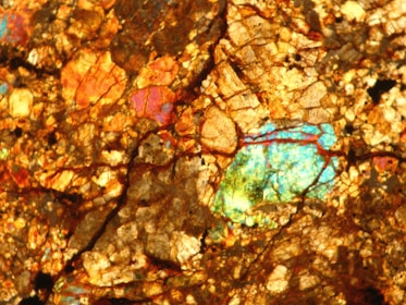 A detailed close-up of a rock or mineral surface, showcasing a vibrant mix of colors and intricate crystalline patterns. The surface features a rich array of gold, orange, and copper hues interspersed with bright patches of blue and green, giving it an iridescent quality.