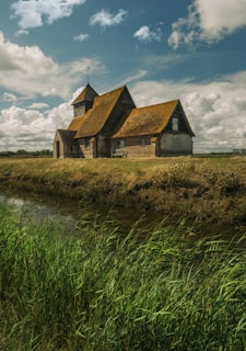 A quaint, rustic church with a traditional design sits on a grassy landscape beside a narrow river. The building features a steep, weathered roof with a small bell tower at the front. Lush green grass and reeds border the water, while fluffy clouds fill the blue sky, casting gentle shadows over the scene.