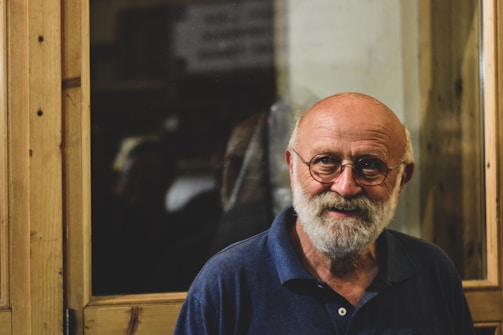 An elderly man with a white beard and round glasses is smiling warmly. He is wearing a navy blue polo shirt. The background features a large wooden window frame and a dimly lit interior.