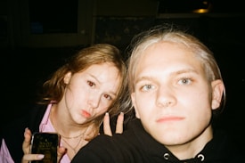 A close-up of two people taking a selfie indoors. The person on the left has long brown hair and is making a duck face, holding a smartphone that shows the time 22:07. The person on the right has blonde hair and is looking directly at the camera. Both are wearing casual clothing.