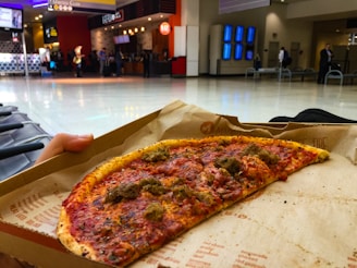 Half-eaten pizza slice on parchment paper in an airport terminal with people and food concessions in the background.