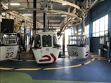 A cable car station with several gondolas waiting to be deployed. The gondolas are numbered and have signage that indicates they belong to the Teleférico in Madrid. The station has a modern industrial look with metal beams and railings. A person is visible inside one of the gondolas, and the floor has colorful markings with a combination of green, yellow, and blue.
