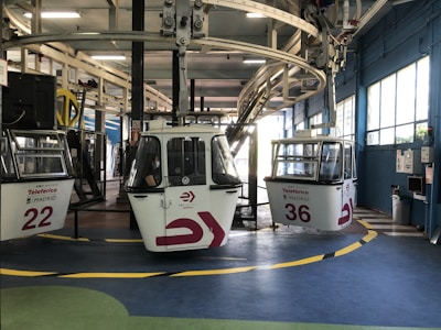 A cable car station with several gondolas waiting to be deployed. The gondolas are numbered and have signage that indicates they belong to the Teleférico in Madrid. The station has a modern industrial look with metal beams and railings. A person is visible inside one of the gondolas, and the floor has colorful markings with a combination of green, yellow, and blue.