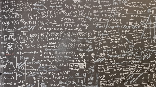 A large blackboard filled with complex mathematical equations, symbols, and annotations written in chalk. The writing is dense and covers the entire surface, indicating a thorough and detailed exploration of mathematical concepts. Various mathematical operations, symbols like integral, summation, and square roots, are visible along with arrows and diagrams.