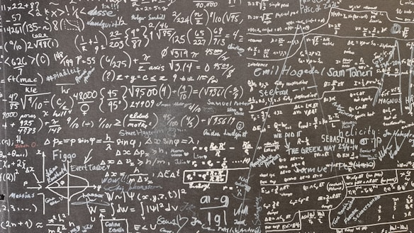 A large blackboard filled with complex mathematical equations, symbols, and annotations written in chalk. The writing is dense and covers the entire surface, indicating a thorough and detailed exploration of mathematical concepts. Various mathematical operations, symbols like integral, summation, and square roots, are visible along with arrows and diagrams.