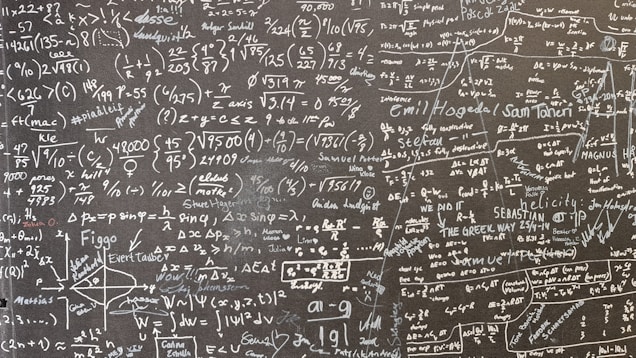 A large blackboard filled with complex mathematical equations, symbols, and annotations written in chalk. The writing is dense and covers the entire surface, indicating a thorough and detailed exploration of mathematical concepts. Various mathematical operations, symbols like integral, summation, and square roots, are visible along with arrows and diagrams.