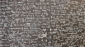 A large blackboard filled with complex mathematical equations, symbols, and annotations written in chalk. The writing is dense and covers the entire surface, indicating a thorough and detailed exploration of mathematical concepts. Various mathematical operations, symbols like integral, summation, and square roots, are visible along with arrows and diagrams.