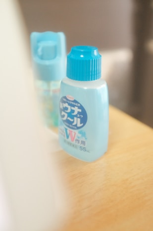 Two small bottles with blue caps sit on a light wooden surface. The bottles appear to be personal care or cosmetic items, displaying labels with Japanese text.