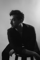 A black and white portrait of a person sitting on a stool, gazing off to the side. The lighting creates soft shadows, highlighting the contours of the face and hairstyle. The background is blurred and plain, putting the focus on the subject. The person is wearing a dark jacket, adding to the classic and timeless feel.