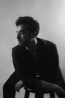 A black and white portrait of a person sitting on a stool, gazing off to the side. The lighting creates soft shadows, highlighting the contours of the face and hairstyle. The background is blurred and plain, putting the focus on the subject. The person is wearing a dark jacket, adding to the classic and timeless feel.