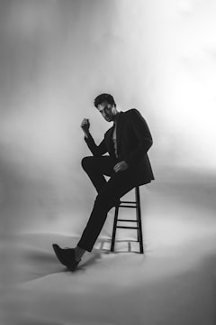 A man dressed in a suit is seated on a stool, positioned in a dimly lit studio setting with a high-contrast, black and white motif. His pose is relaxed and casual, with one foot touching the ground and the other slightly elevated, creating a dynamic, diagonally oriented line. The lighting creates soft shadows around him, enhancing the mood and highlighting the texture of the suit.