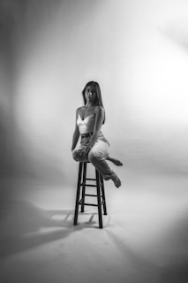 A woman is seated on a tall stool in a minimalist studio setting. She wears a fitted top and jeans, with her long hair draping over one shoulder. The backdrop is smooth and expansive, with soft lighting casting gentle shadows.