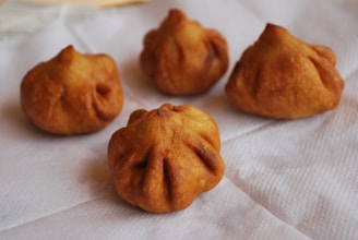 Four fried pastries are placed on a white paper surface. They have a golden-brown color with a distinctive folded pattern, resembling dumplings or puffs.