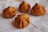 Four fried pastries are placed on a white paper surface. They have a golden-brown color with a distinctive folded pattern, resembling dumplings or puffs.