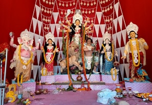 A vibrant and colorful display of Hindu deities, including a central figure of Goddess Durga adorned with intricate decorations and ornaments. She is depicted riding a lion with several arms holding various weapons. Surrounding her are other deities, including an elephant-headed figure, possibly Lord Ganesha, to the left. The backdrop is a bright red with geometric patterns and hanging decorations. The setup has offerings and decorative items placed in front.