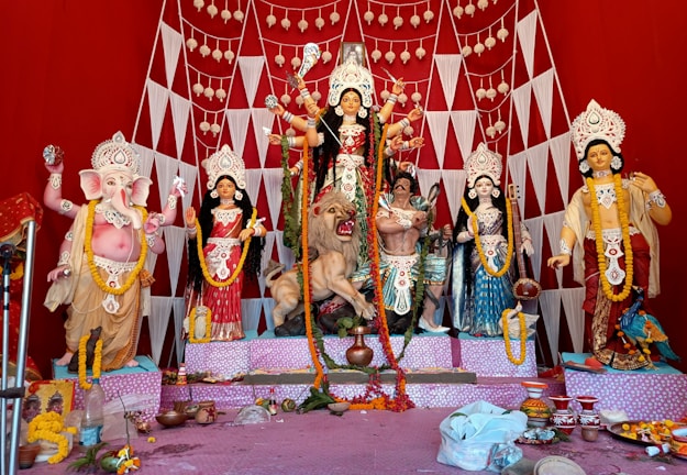 A vibrant and colorful display of Hindu deities, including a central figure of Goddess Durga adorned with intricate decorations and ornaments. She is depicted riding a lion with several arms holding various weapons. Surrounding her are other deities, including an elephant-headed figure, possibly Lord Ganesha, to the left. The backdrop is a bright red with geometric patterns and hanging decorations. The setup has offerings and decorative items placed in front.