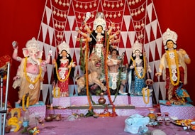 A vibrant and colorful display of Hindu deities, including a central figure of Goddess Durga adorned with intricate decorations and ornaments. She is depicted riding a lion with several arms holding various weapons. Surrounding her are other deities, including an elephant-headed figure, possibly Lord Ganesha, to the left. The backdrop is a bright red with geometric patterns and hanging decorations. The setup has offerings and decorative items placed in front.
