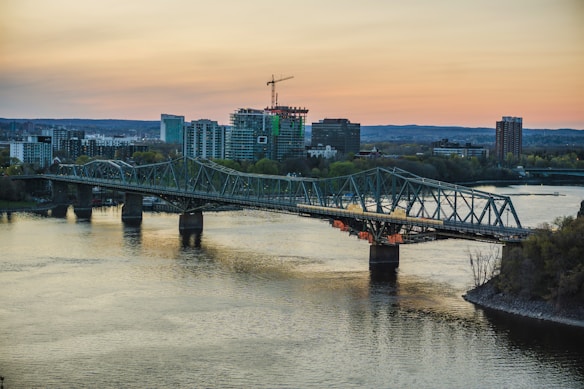 A steel bridge stretches across a wide river, with reflections visible in the water. The horizon features a cityscape of modern buildings under a pastel sky during sunset, where some construction is evident with a crane visible atop a building.
