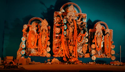 A display of intricately crafted religious statues adorned with ornate jewelry and traditional clothing, surrounded by decorations. The central figure appears larger and more elaborately decorated, with others flanking on either side, all set against a turquoise backdrop.