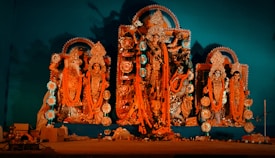 A display of intricately crafted religious statues adorned with ornate jewelry and traditional clothing, surrounded by decorations. The central figure appears larger and more elaborately decorated, with others flanking on either side, all set against a turquoise backdrop.