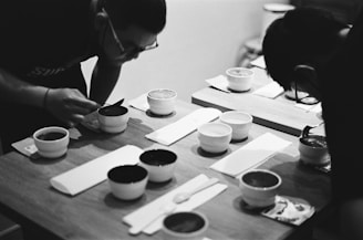 Two individuals are leaning over a wooden table examining or tasting the contents of small white bowls. The setting appears intimate and focused, with a monochrome aesthetic adding a classic and timeless feel. Utensils are neatly arranged beside the bowls.