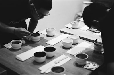 Two individuals are leaning over a wooden table examining or tasting the contents of small white bowls. The setting appears intimate and focused, with a monochrome aesthetic adding a classic and timeless feel. Utensils are neatly arranged beside the bowls.