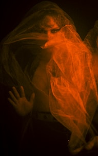 A person is partially obscured by thin, flowing, orange translucent fabric that creates a mysterious and ethereal effect. The soft lighting casts shadows, adding depth and drama. One hand of the person is visible, slightly pressed against an unseen surface, and their face is partially hidden behind the fabric, emphasizing their eyes.