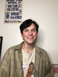 A person with dark hair is smiling, wearing a plaid shirt over a printed t-shirt. On the wall behind, there is a vintage wrestling poster with blue text featuring names like Watson, Valentine, Kiniski, and Von Erich. The setting appears casual and relaxed.