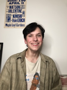 A person with dark hair is smiling, wearing a plaid shirt over a printed t-shirt. On the wall behind, there is a vintage wrestling poster with blue text featuring names like Watson, Valentine, Kiniski, and Von Erich. The setting appears casual and relaxed.
