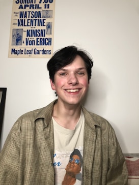 A person with dark hair is smiling, wearing a plaid shirt over a printed t-shirt. On the wall behind, there is a vintage wrestling poster with blue text featuring names like Watson, Valentine, Kiniski, and Von Erich. The setting appears casual and relaxed.