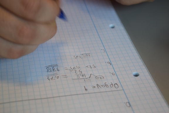 A close-up of a hand holding a pen, writing mathematical calculations on a sheet of graph paper. The paper is blue and white, filled with numbers in a column format, possibly performing division or multiplication tasks.