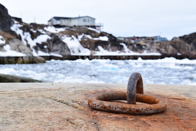 A rusted metal ring is embedded in a rough, weathered concrete surface in the foreground. In the background, snow-dusted rocky terrain is visible along with a choppy, icy body of water. A modest house with muted colors is perched on the hill, and the overall setting suggests a cold, rugged coastal environment.