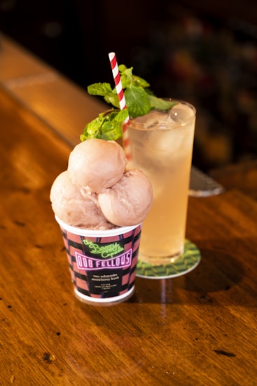 A wooden surface holds a cup with three scoops of ice cream and a cold beverage in a tall glass. The ice cream cup features a distinctive design with a red and blue pattern and a readable label. A red and white striped straw sticks out of the ice cream, accented with a sprig of mint. The glass is filled with a light-colored drink and ice cubes, sitting on a green coaster.
