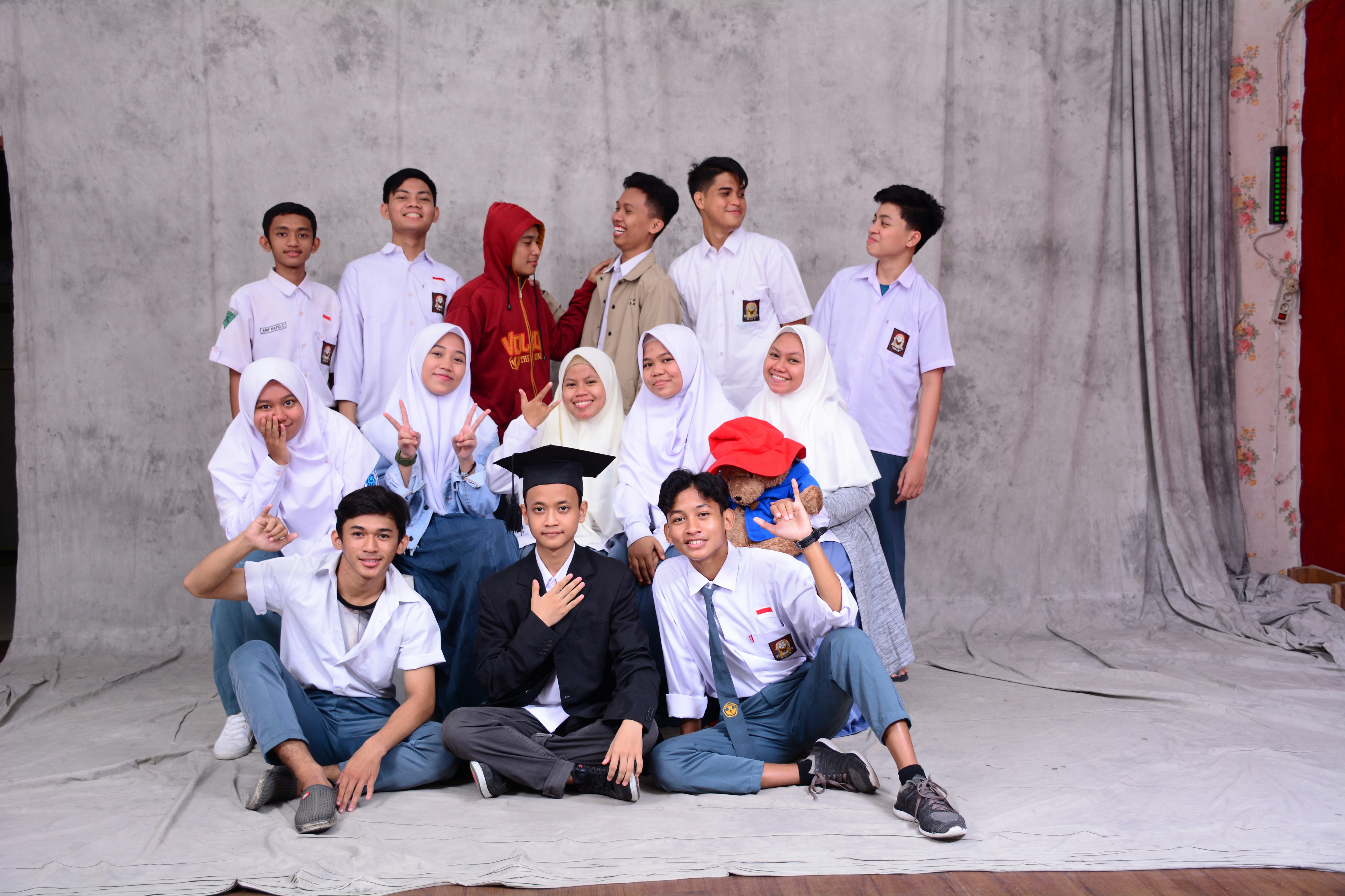 A group of students, mostly wearing school uniforms, pose for a picture against a plain backdrop. Some are seated in front while others stand behind. The students show a range of expressions from smiles to playful gestures. There is a person in the center wearing a graduation cap, adding a celebratory tone.