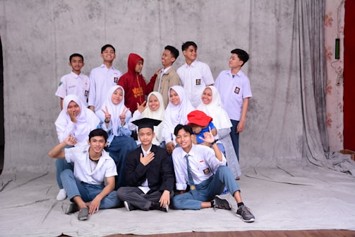 A group of students, mostly wearing school uniforms, pose for a picture against a plain backdrop. Some are seated in front while others stand behind. The students show a range of expressions from smiles to playful gestures. There is a person in the center wearing a graduation cap, adding a celebratory tone.