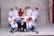 A group of students, mostly wearing school uniforms, pose for a picture against a plain backdrop. Some are seated in front while others stand behind. The students show a range of expressions from smiles to playful gestures. There is a person in the center wearing a graduation cap, adding a celebratory tone.
