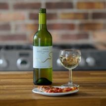 A bottle of Pinot Grigio wine is placed next to a glass filled with the wine on a wooden table. In front of them, a small white plate holds a slice of pizza. The background features a blurred view of a stovetop and a brick wall, creating a cozy kitchen ambiance.