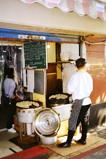 A street food vendor is seen cooking food on large, round griddles. The vendor is dressed in a white shirt with black striped apron and a hat. Another person standing inside the shop can be seen in the background. A board with menu items written in a foreign language is hanging above the cooking area.