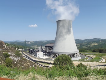 A large cooling tower is prominently visible, emitting steam against a clear blue sky. The landscape surrounding it is mountainous with patches of greenery and sparse vegetation. The power facility has several industrial structures and pipelines leading to the tower, indicating an industrial or energy generation site.